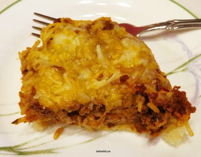 Slice of deep dish taco casserole with seasoned turkey, cheese, refried beans, and taco rice on pizza crust