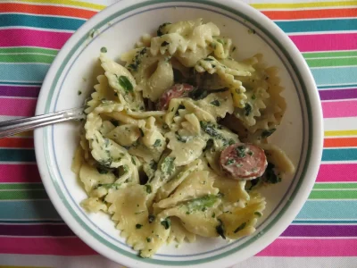 Close-up of a single serving bowl of leftover pasta containing bowtie noodles, Alfredo sauce, spinach, and smoked sausage.