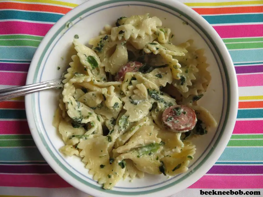 Close-up of a single serving bowl of leftover pasta containing bowtie noodles, Alfredo sauce, spinach, and smoked sausage.