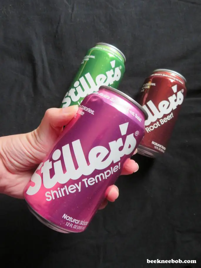 close-up of a hand holding a can of Stiller's Shirley Temple soda with the other two flavors positioned in the background.