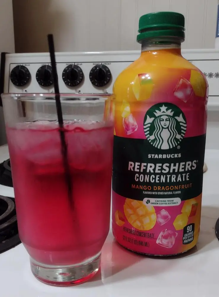 A bottle of Starbucks Refreshers Concentrate in Mango Dragonfruit next to a glass of the prepared pink beverage.