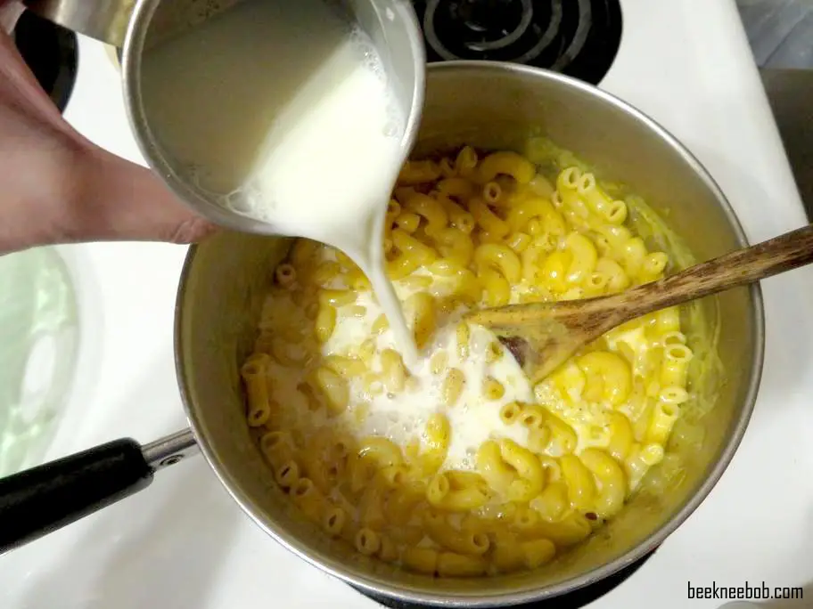 a cup of milk pouring into a large saucepan of prepared macaroni and cheese on the stove for a baked macaroni and cheese recipe